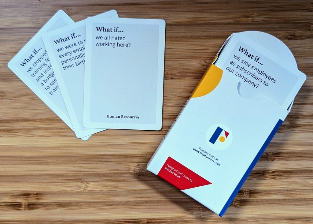 Image contains three playing cards and a box. Only the text on the first card is visible and reads "what if we all hated working here?" There is a card poking out the box which reads "What if... we saw employees as subscribers to our company?"  The box is white with the Freeformers logo.